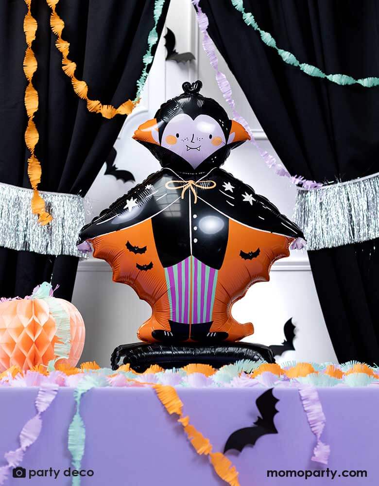 A festive Halloween party featuring Momo Part's 31" x 44" Dracula shaped standing foil balloon in black and orange on a purple party table. Around the balloon are colorful party streamers, a silver tinsel garland and orange pumpkin honeycomb on the table with bat paper decorations. Makes it a cute party inspiration for kid's not so scary Halloween party.