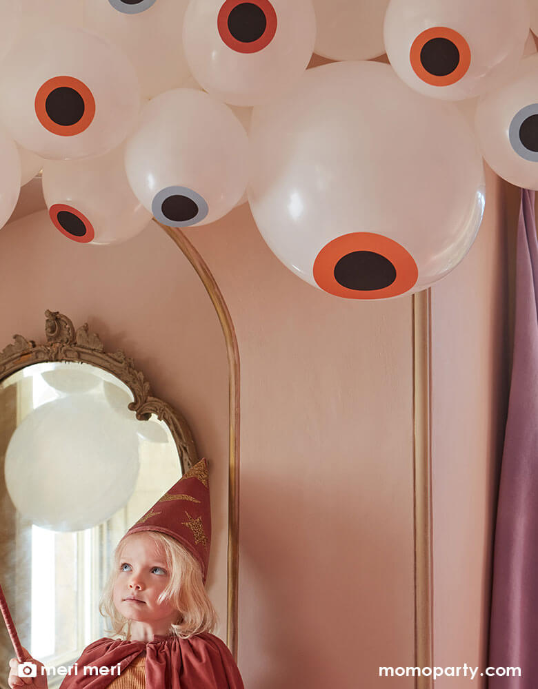 A girl in her witch costume with a wand looking at Momo Party's Eyeball Balloon Garland by Meri Meri hung above the ceiling in a pink room. Makes it a whimsical inspiration for kid's not-so-scary Halloween celebrations.