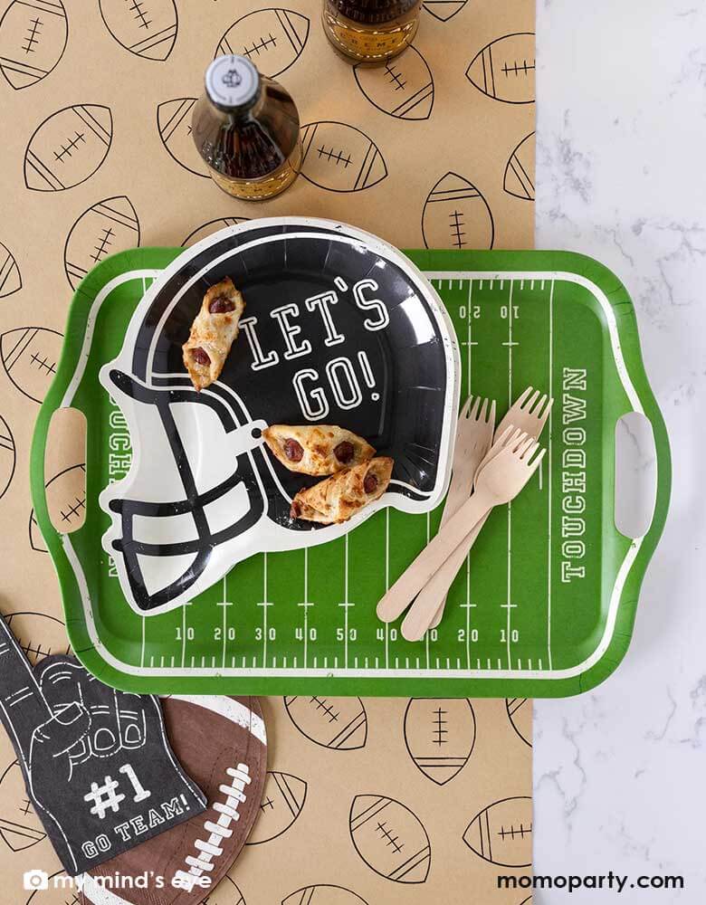 A football party table features Momo Party's Football Field bamboo reusable tray, on the tray it's a black helmet shaped plate featuring "Let's Go"" on it, no the plates there some party snacks and around the tray there are Momo Party's football shaped napkins and #1 finger shaped black napkin on a light brown table runner with football illustration on it. With party drinks around, it makes a great party table decoration for a fun football season watch party!