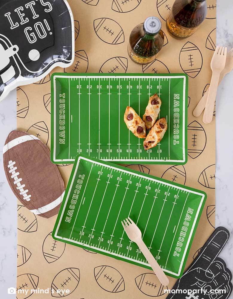 A football party table features Momo Party's Football Field shaped plates, on the plate there are some party snacks and around the plates there are Momo Party's football shaped napkins, black helmet shaped plates featuring "Let's Go"" on it, and a no #1 finger shaped black napkin on a light brown table runner with football illustration on it. With party drinks around, it makes a great party table decoration for a fun football season watch game party or a Super Bowl party!