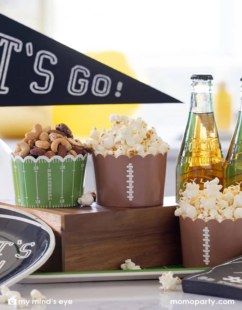 A football themed party table featuring Momo Party's jumbo football food cups filled with watching party snacks including popcorn and mixed nuts. Next to them is some root beer bottles and "Let's Go" felt party pennant by My Mind's Eye.