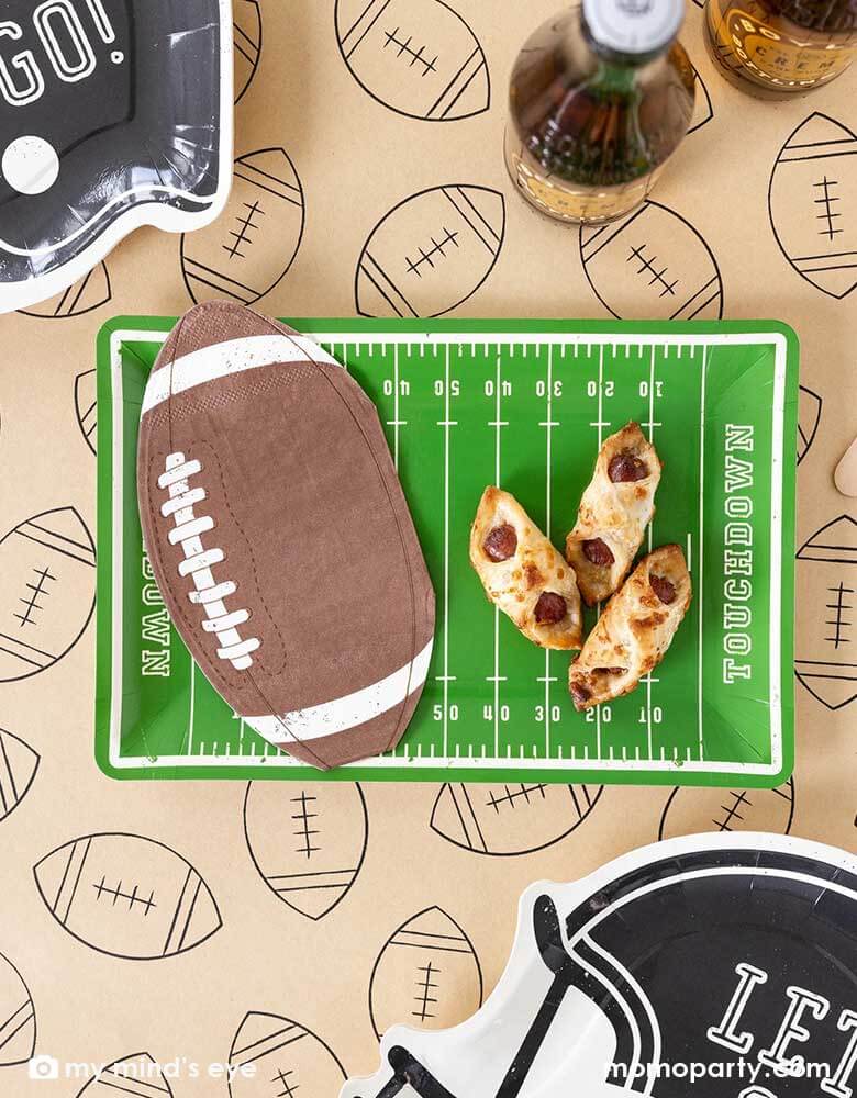 A football party table features Momo Party's Football Field shaped plate, on the plate there are some party snacks and some football shaped napkins, around the plates there are Momo Party's football shaped black helmet shaped plates featuring "Let's Go"" on them. All these are on a light brown table runner with football illustrations on it. With party drinks around, it makes a great party table decoration for a fun football season watch game party or a Super Bowl party!