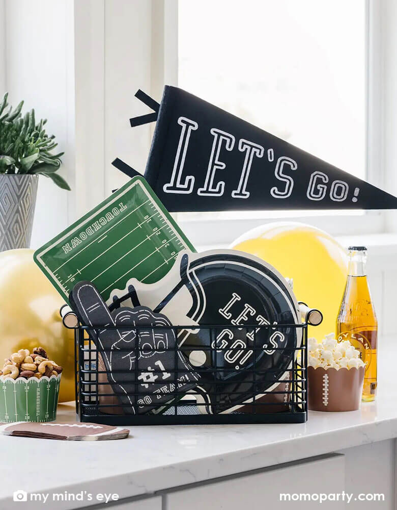An occasion basket filled with Momo Party's football themed party decorations including a "Let's Go" felt party pennant, football field shaped serving plates, black football helmet shaped plates, black #1 finger shaped napkins and football themed foodcups filled with watching party snacks including popcorn and nuts, next to it are some yellow party balloons and some root beer bottles. Perfect for a watching game, super bowl party in football season!