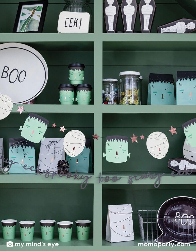 A green shelf filled with Momo Party's Halloween party supplies including Frankenstein and mummy themed plates, cups, goodie bags, treat boxes and napkins. In the front of the shelf, there's a Frank & Mummy garland hung, featuring spooky best friends, copper foiled stars, and a stitched word banner, these banners a sure to be a frightful delight at your Halloween parties!