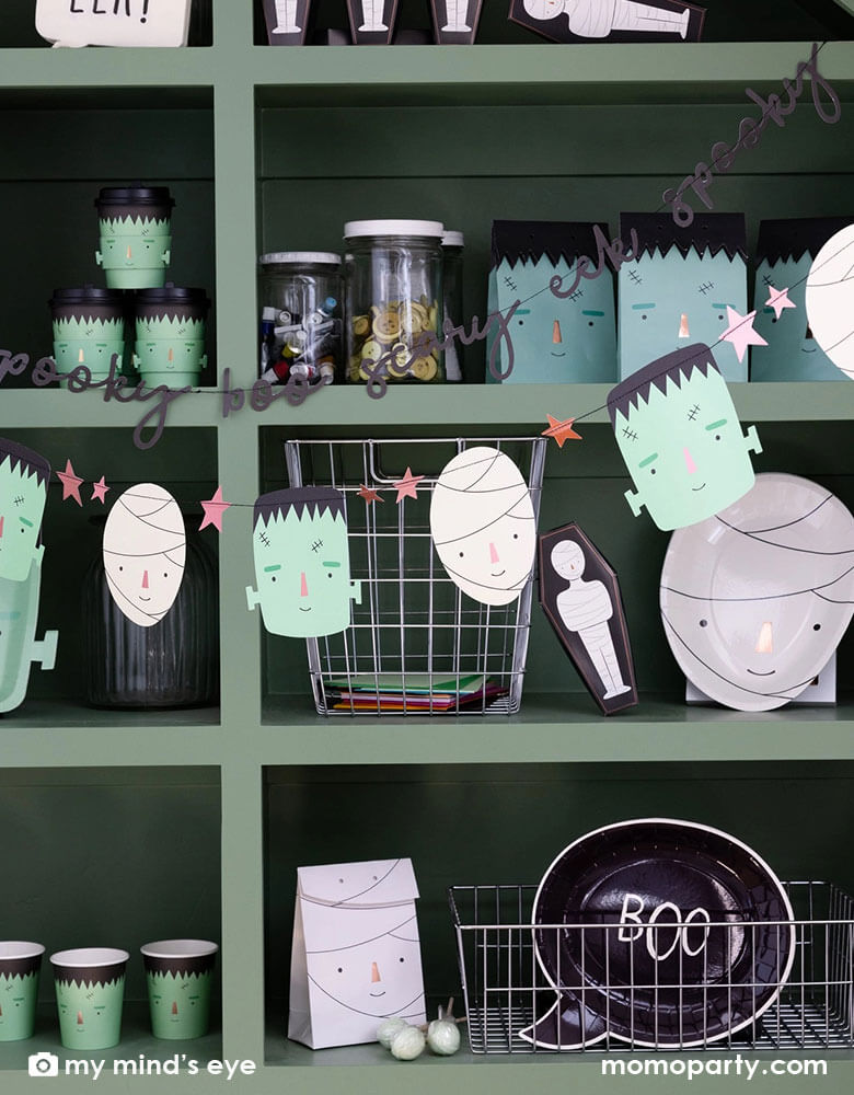 A green display shelf filled with Momo Party's Halloween party supplies including Frankenstein and mummy themed plates, cups, goodie bags, treat boxes and napkins. In the front of the shelf, there's a Frank & Mummy garland hung,  featuring spooky best friends, copper foiled stars, and a stitched word banner, these banners a sure to be a frightful delight at your Halloween parties!