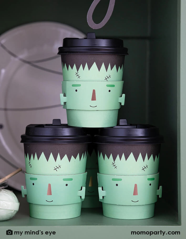 Three Momo Party's 8 oz Frank and Mummy Cozy to-go cups by My Mind's Eye stacked together on a shelf. Featuring a friendly Frankenstein sleeve these cozy to go cups will keep your favorite beverage warm through all the tricks and treats on Halloween night!