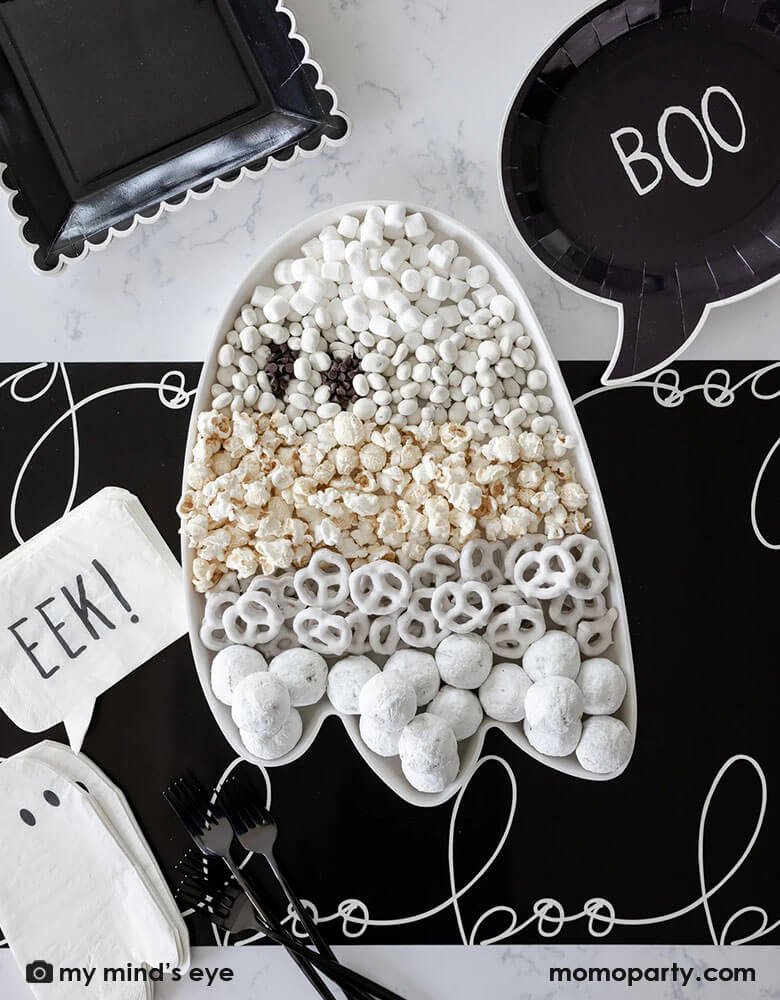 A black and white Halloween table featuring Momo Party's 12" x 16" ghost shaped bamboo reusable tray, filled with all sorts of Halloween treats and goodies including white donut holes, white sugar coated pretzels, popcorn, white chocolates and marsh mellow. Around it it's Momo Party's black boo shaped plates, ghost shaped napkins with EEK! shaped napkins on a black table runner with "boo" scripted font written over it. All makes a cool and spooky ideas for a kid's Halloween party table setting inspiration.