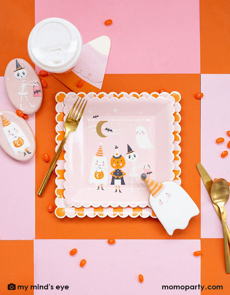 A pink Halloween party table with orange and pink checkered tablecover, featuring Momo Party's ghoul gang collection by My Mind's Eye including ghoul gang scene scallop edged square plates and orange and pink checkered scallop edged plates. With coordinating decorated Halloween sugar cookies in the adorable colors of light pink and orange with orange jelly beans around the table, it makes an adorable pink girly Halloween party inspiration.