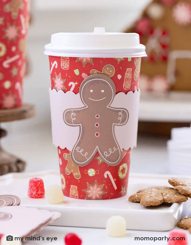A gingerbread house decoration party featuring Momo Party's 16 oz Red Gingerbread Man To-Go Cups by My Mind's Eye. In the back of the table you can see a gingerbread house decorated with peppermint and candies, plus a treat bucket with gingerbread man pattern design filled with festive gingerbread cookies and candies.