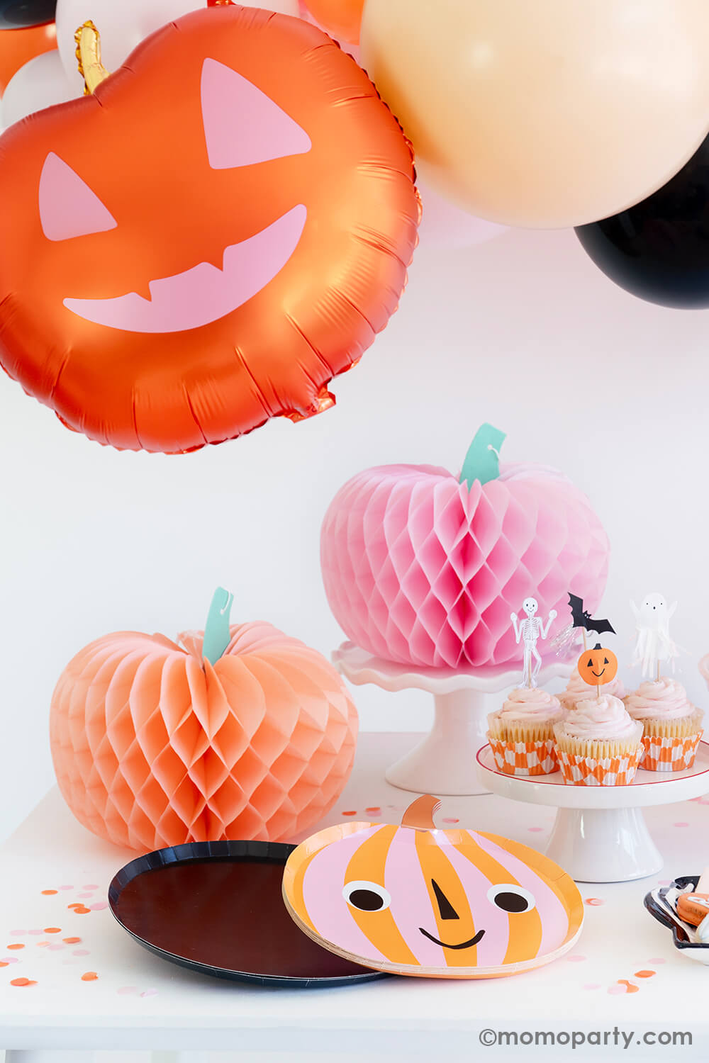 Momo Party's "Hey Pumpkin" party box featuring two honeycomb pumpkins in boo-tiful colors of pink and peach. With a smiley pumpkin shaped plates and a set of cute cupcakes topped with Halloween character toppers including a pumpkin, a skeleton, a bat and a ghost, with a happy pumpkin foil balloon hung above the table, it creates a spooky yet cute vibe for a fun Halloween party.