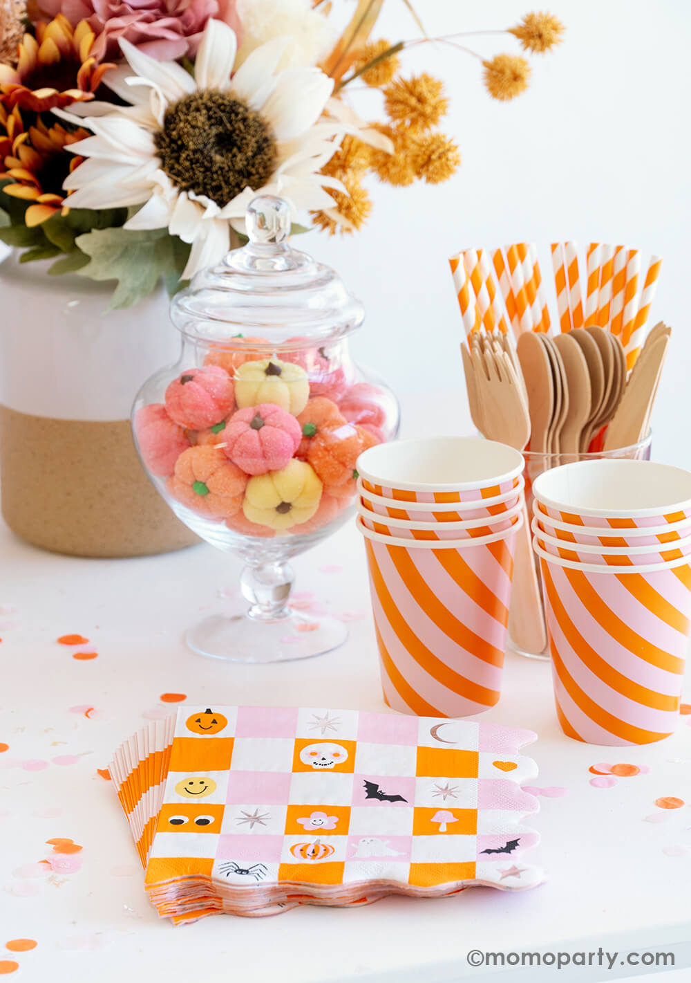 A beautiful Halloween party table featuring groovy Halloween checkered napkins with skull, pumpkin, bat, moon characters on them . Next to the napkins are some orange/pink party cups next to a candy jar filled with pumpkin marshmallow in the colors of orange, pink and light yellow. In the back is a fall flower arrangement that set an autumn theme for a halloween party.
