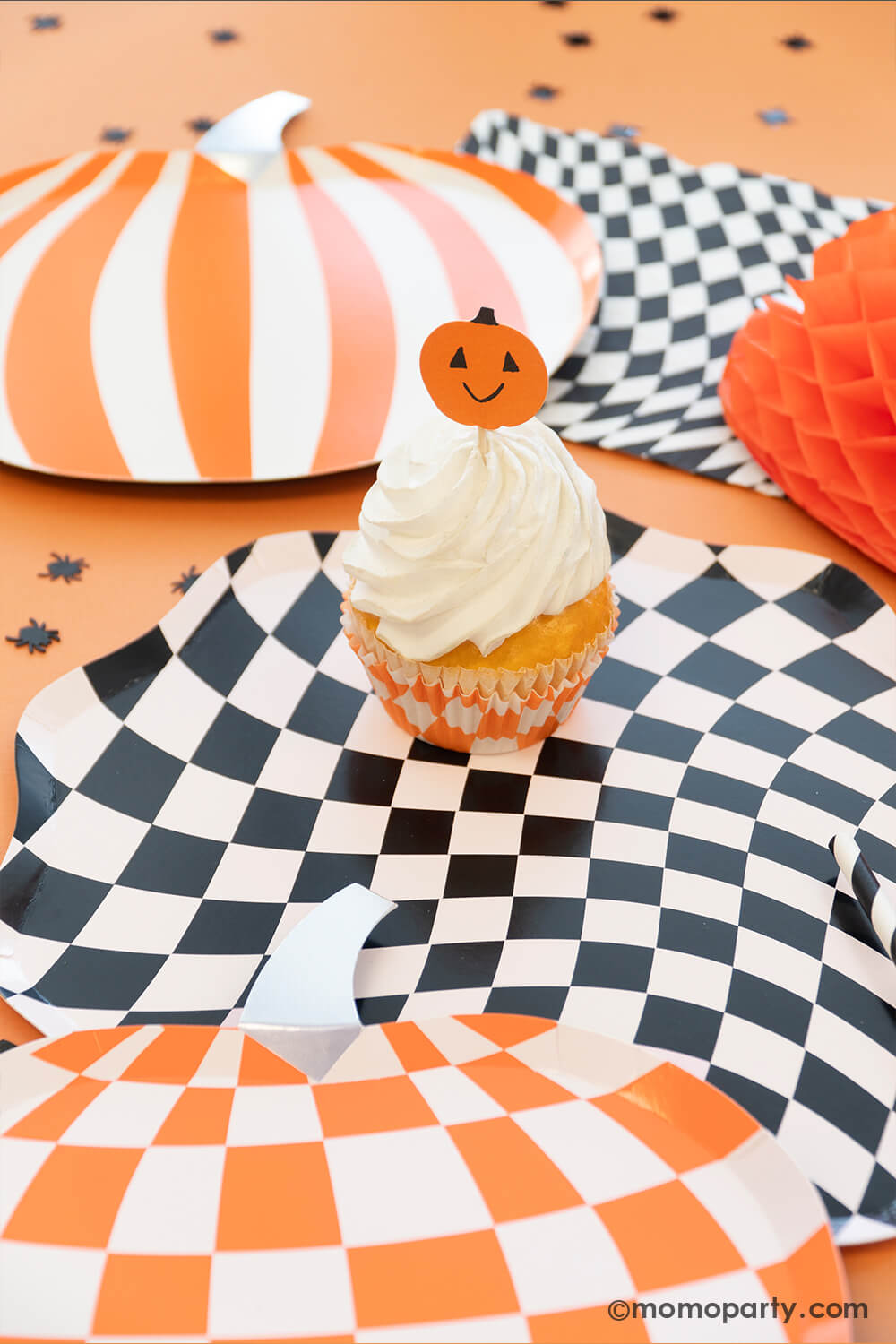A festive Halloween party table featuring Momo Party's Halloween swirl checkered plates, napkins and cups, along with mod patterned pumpkin shaped plates in orange and black, with a honeycomb spider decoration and confetti around it makes a spooky yet fun inspiration for a kid's friendly Halloween party.
