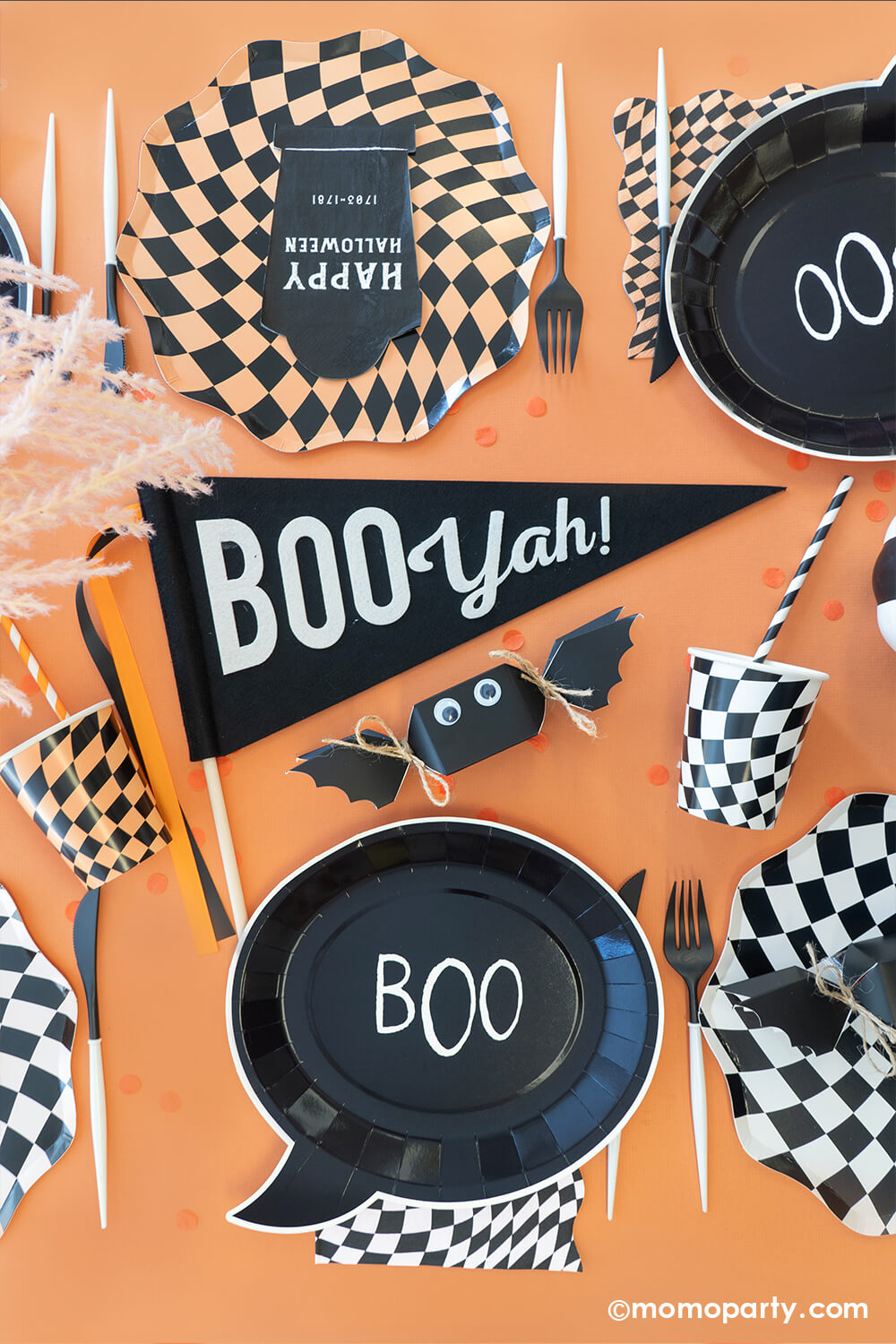 A table filled with Momo Party Classic Orange and Black Halloween Party Decorations including swirling checkered plates, cups, napkins, Boo Yah party pennant, bat shaped jelly beans, black tombstone shaped napkins and boo shaped plates, all makes a spooky yet fun decoration ideas for a Halloween bash this season.