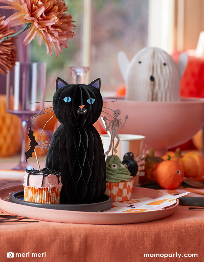 A pink and orange Halloween party table decorated with Momo Party's Halloween black cat and ghost honeycomb decorations by Meri Meri and a variety of Halloween themed tableware and cupcake toppers in the design of bats, skeletons, pumpkins and spiders. Makes it a whimsical inspiration for kid's not-so-spooky Halloween party or celebration.