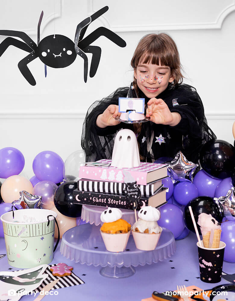 A girl playing with Momo Party's Halloween Guessing Game Set by Party Deco at a Halloween party table decorated with a purple tablecloth and black balloons. On the table there are some Halloween themed treats including ghost cupcakes, pink cotton candy, Halloween sugar cookies on Momo Party's pink skull shaped plates. Above the party table there's an adorable smiley spider paper decoration hung above, makes it a cute inspiration for kid-friendly Halloween party set up.