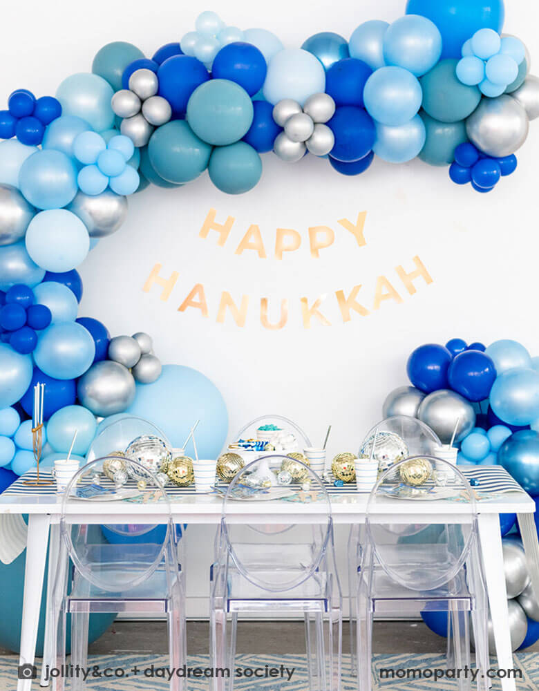 A festive Hanukkah party table set up featuring a large blue balloon garland and "Happy Hanukkah" letter banner above the party table. On the table are Momo Party's blue striped plates and party cups with Festival of Lights small star shaped plates and patterned napkins by Daydream Society. On the blue striped table runner are some silver and gold disco balls in different sizes and Chanukah candles. All makes it a great inspo for a festive Hanukkah party for family gathering.