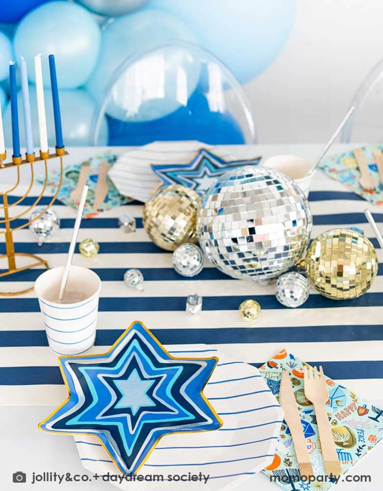 A festive Hanukkah party table featuring Momo Party's blue striped plates and party cups with Festival of Lights small star shaped plates and patterned napkins by Daydream Society. On the blue striped table runner are some silver and gold disco balls in different sizes and Chanukah candles. In the back you can see a blue balloon garland in different shades of blue, makes it a great inspo for a festive Hanukkah party for family gathering.