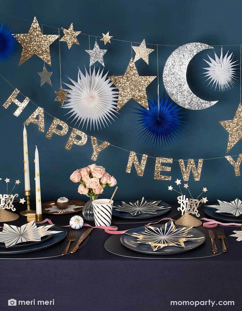 A festive New Year Party Table featuring Momo Party's gold star striped plates and napkins, gold striped party cups and Happy New Year party hats on a dark navy covered table. Above the table hung Momo Party's 8 ft New Year party garland with gold and silver glittered moon and star pennants, along with navy and white paper fans, below it is a gold glittered Happy New Year letter banner, makes it a great inspiration for a festive yet elegant New Year countdown party.