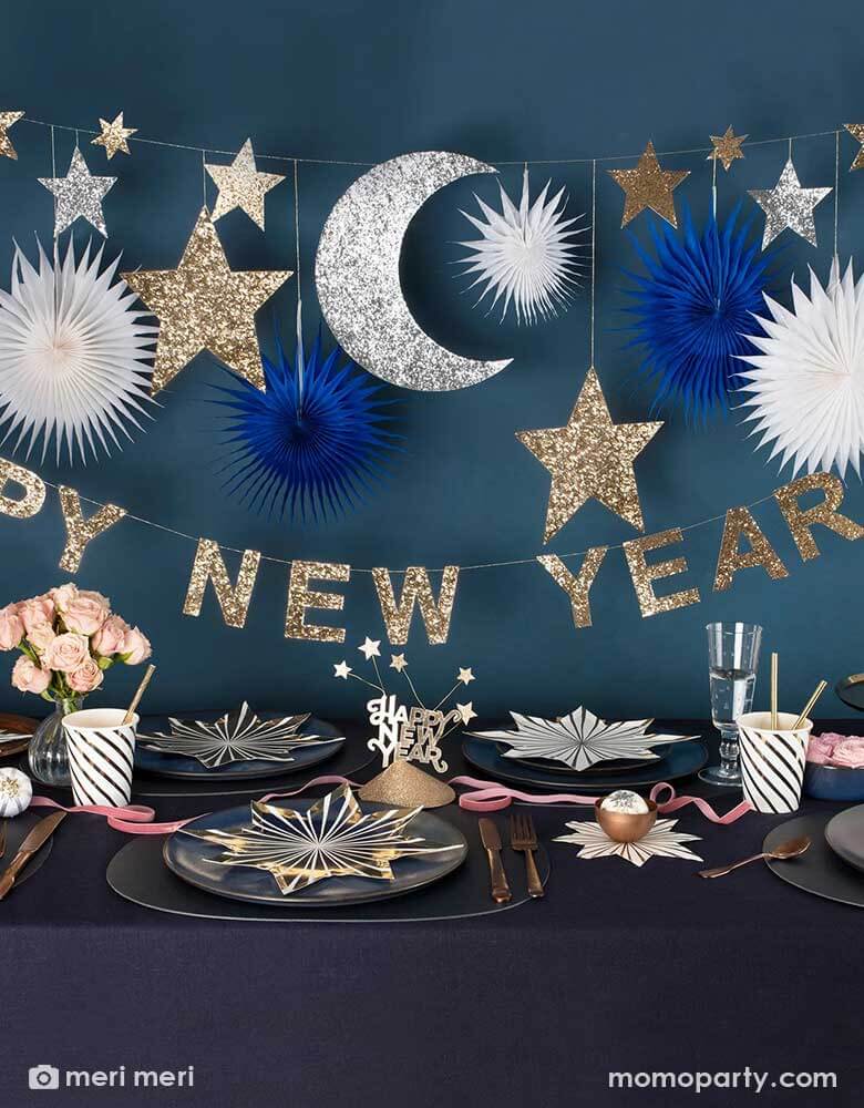 A festive New Year Party Table featuring Momo Party's gold star striped plates and napkins, gold striped party cups and Happy New Year party hats on a dark navy covered table. Above the table hung Momo Party's 8 ft New Year party garland with gold and silver glittered moon and star pennants, along with navy and white paper fans, below it is a gold glittered Happy New Year letter banner, makes it a great inspiration for a festive yet elegant New Year countdown party.