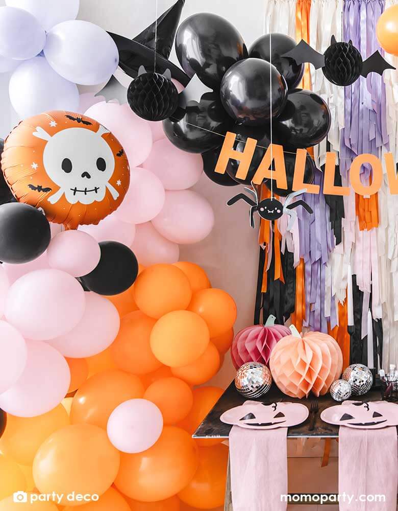 A festive Halloween party set up featuring a beautiful balloon garland in orange, pink, lilac and black, with Momo Party's 14" orange round skull foil balloon, witch's hat, paper spider hanging decoration and honeycomb bats in front of a wall of colorful fringe in the Halloween themed colors. In the front there's a party table with some honeycomb pumpkins, disco balls and pink pumpkin shaped plates. All makes this a perfect party inspiration for a not so spooky kid's friendly Halloween party.