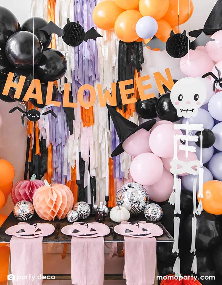 A festive Halloween party set up featuring a beautiful balloon garland in orange, pink, lilac and black, with Momo Party's 14" orange round skull foil balloon, witch's hats, skeleton hanging decoration, paper spider hanging decoration and honeycomb bats in front of a wall of colorful fringe in the Halloween themed colors. In the front there's a party table with some honeycomb pumpkins, disco balls and pink pumpkin shaped plates. 