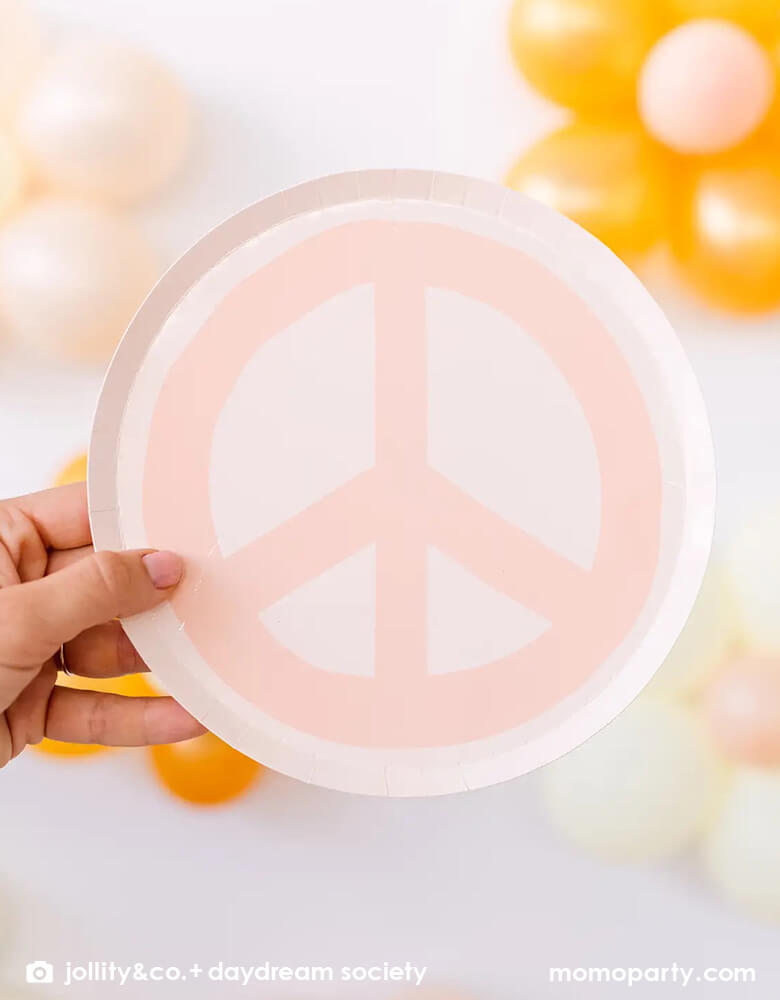A hand holding Momo Party's 8" peach love and peace sign dessert plate by Jollity & Co. in front of a wall decorated with daisy balloons. Momo Party's 8" love and peace smily face dessert plate by Jollity & Co. Featuring a groovy, retro design and bursting with boho vibes, these plates will bring good vibes to any girl's birthday.