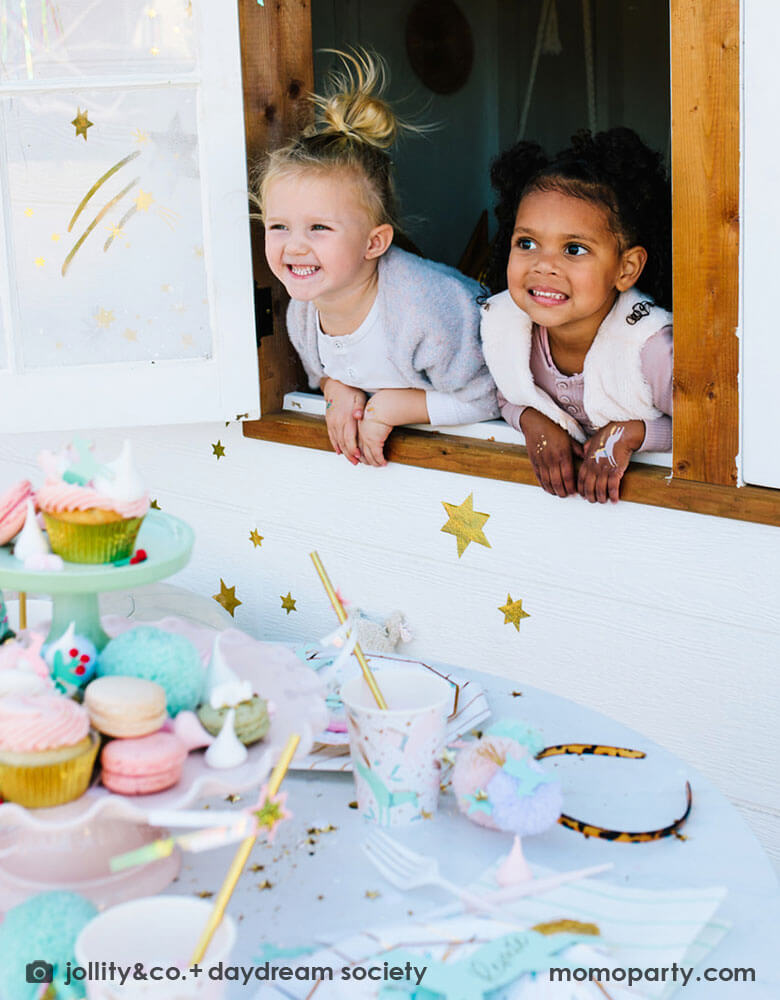 Two school aged girls leaning against a window looking out a party table featuring Momo Party's magical unicorn partyware including plates and cups. In the middle of the table, a cupcake tower with a sorts of treats including cupcakes, macarons, cotton candies and more, makes this a whimsical Christmas holiday set up that's perfect for kids.
