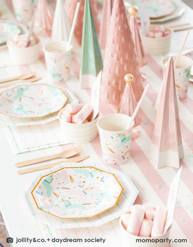 A pink Christmas party table featuring Momo Party's magical unicorn plates and cups by Daydream Society. A few ceramic Christmas tree decorations in mint and pink are used as the centerpiece on a pink white striped table runner. On the table you can see some pink marsh mellows in food cups as the treats for kids.