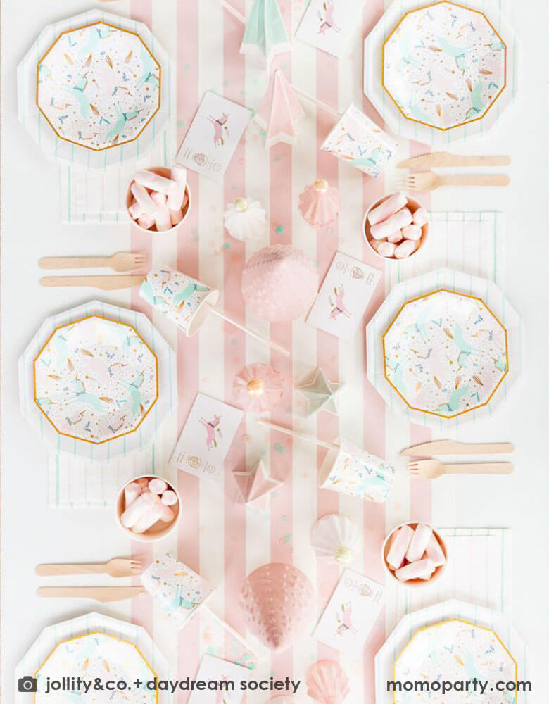A pink Christmas party table featuring Momo Party's magical unicorn plates and cups by Daydream Society. A few ceramic Christmas tree decorations in mint and pink are used as the centerpiece on a pink white striped table runner. On the table you can see some pink marsh mellows in food cups as the treats for kids. All makes this a dreamy whimsical kid's holiday party inspo. 