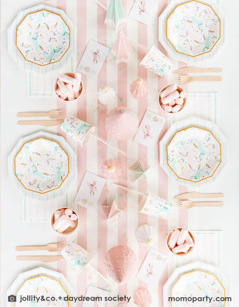 A pink Christmas party table featuring Momo Party's magical unicorn plates and cups by Daydream Society. A few ceramic Christmas tree decorations in mint and pink are used as the centerpiece on a pink white striped table runner. On the table you can see some pink marsh mellows in food cups as the treats for kids. All makes this a dreamy whimsical kid's holiday party inspo.