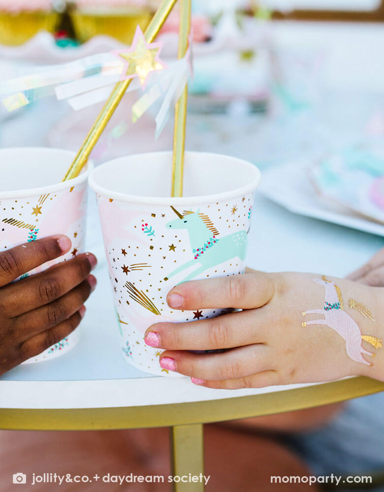 Two girl's hands holding Momo Party's magical unicorn party cups by Daydream Society. On on of the girls' hand there's a temporary tattoo of a unicorn on it. In the party cups, there are gold foil straws decorated with shooting star toppers to create a magical and whimsical vibe for a Christmas Holiday celebration party.