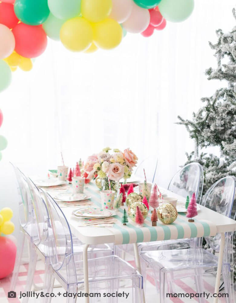 A Christmas table featuring Momo Party's Merry and Bright party collection by Daydream Society. On the table you can see the festive Christmas themed plate, cups and napkins on a mint striped table runner. The middle of the table is decorated with bottle brush Christmas trees in pastel colors. Above the party table, there is a balloon garland in the colors of coral, yellow, pink and mint hung over it. Finally on the right of the table, there's a big Christmas tree covered with frost.