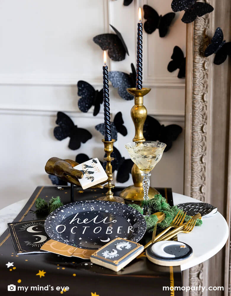 A mystical Halloween table decorated with Momo Party's moons and stars themed party supplies including a crystal ball shaped plate, moons and stars gold foil napkins, moons and stars table runner by My Mind's Eye. With gold metal candle stands holding black candles and vintage gold utensils with Halloween themed sugar cookies and black glittered butterfly decorations on the wall, all makes a perfect inspo for an elegant and chic wizard and witch themed Halloween party decorating idea.