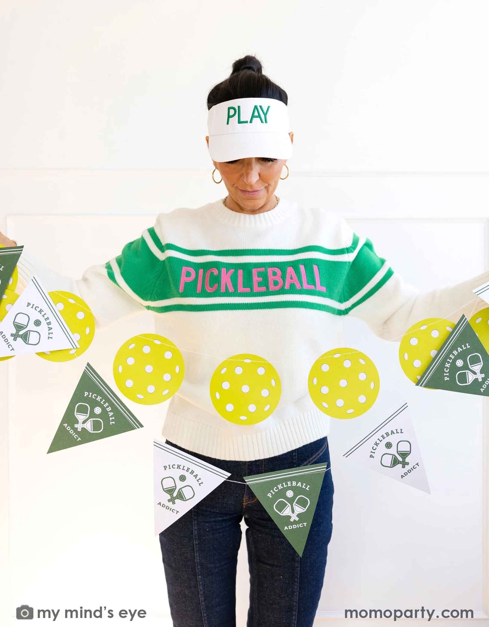 A lady wearing a 'Pickleball' themed sweater and pants with her white hat says "Play" holding Momo Party's pickleball party banner set by My Mind's Eye on a white background.