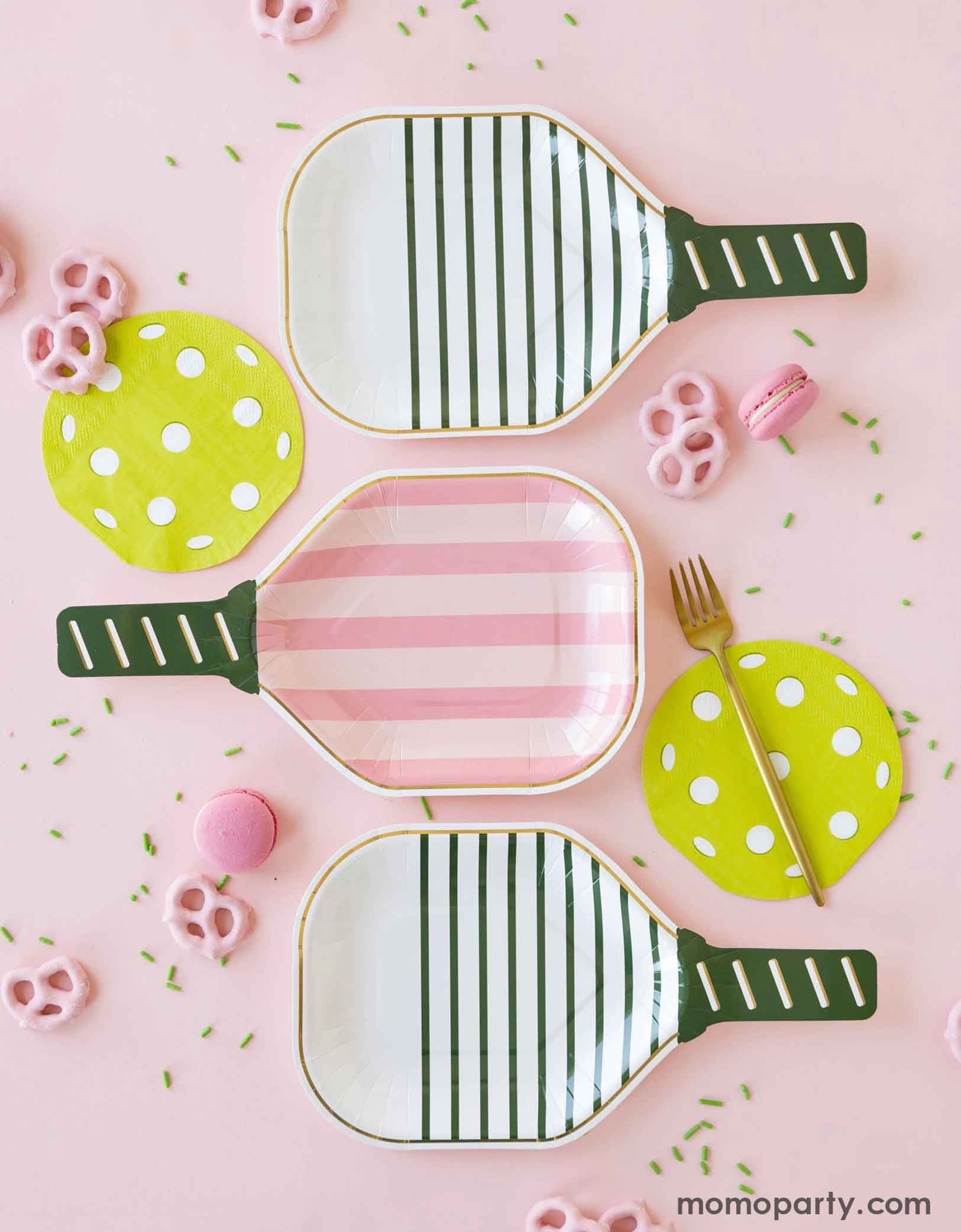 An overhead shot of Momo Party's three pickleball paddle shaped plates in pink, white and green, along with pickleball shaped napkins by My Mind's Eye with pink sprinkles, pink pretzels and macarons around on a pink background. A perfect inspo for decorating a modern and chic paddleball party.
