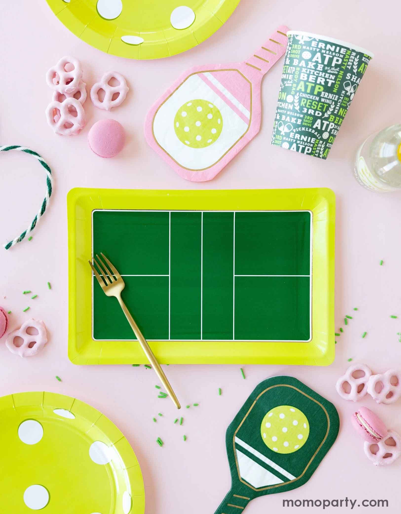 An overhead shot of pink and green table with pickleball-themed party supplies from Momo Party including the pickleball court shaped plate, pickleball shaped plates and pickleball paddle shaped napkins in pink and green and the pickleball party cup by My Mind's Eye on a pink tablecloth. Along with some pink macarons, pretzels and a golden fork - making this an inspiration for a chic and modern pickleball themed celebration.