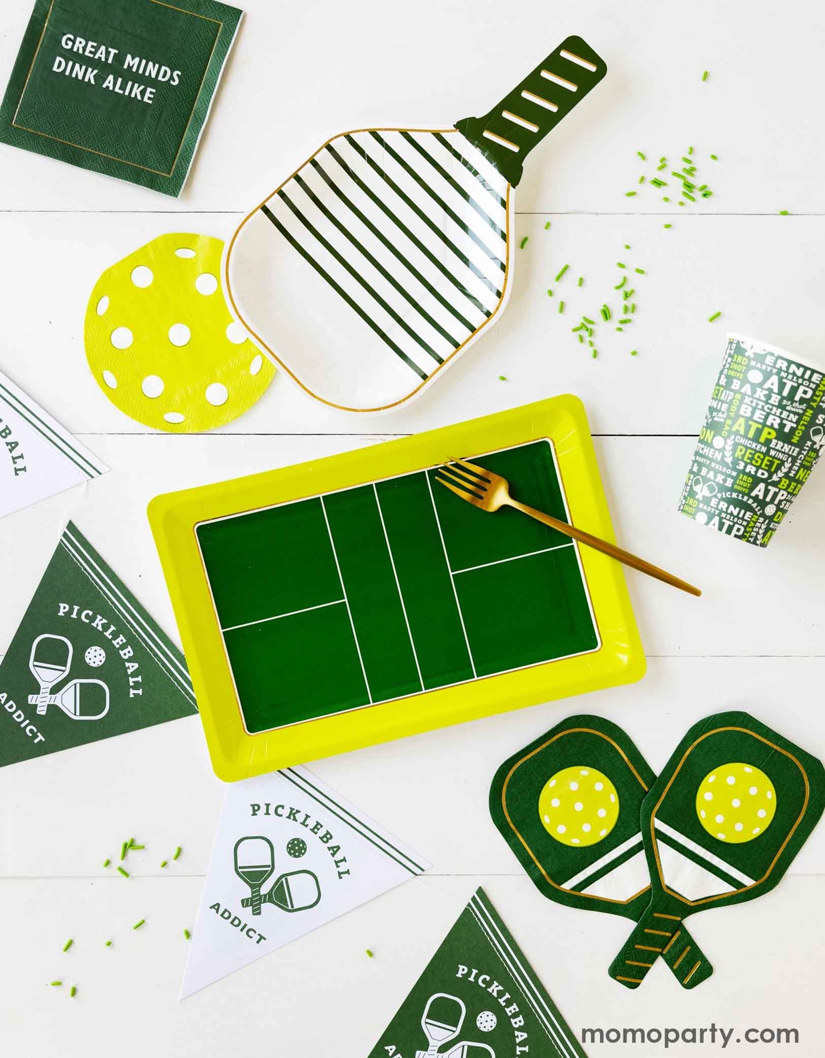 An overhead shot of a table with pickleball-themed party supplies from Momo Party including pickleball court shaped plate, the white pickleball paddle shaped plate, the green pickleball sayings napkins, the pickleball shaped napkin and green paddle shaped napkins, the pickleball party cup and the white and green pickleball party banner by My Mind's Eye. Along with some green sprinkles spread across the table - making this an inspiration for a modern and chic pickleball themed celebration.