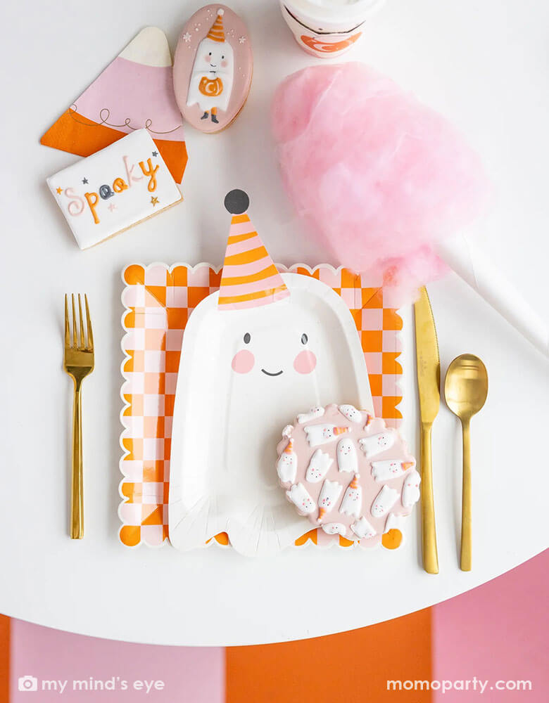 A pink Halloween party table featuring Momo Party's ghoul gang collection by My Mind's Eye including 11.5" x 6.5" ghost shaped plates, orange and pink checkered scallop edged plates and ghoul gang pink to-go cups. With coordinating decorated sugar cookies and pink cotton candy around the table, it makes an adorable pink girly Halloween party inspiration.