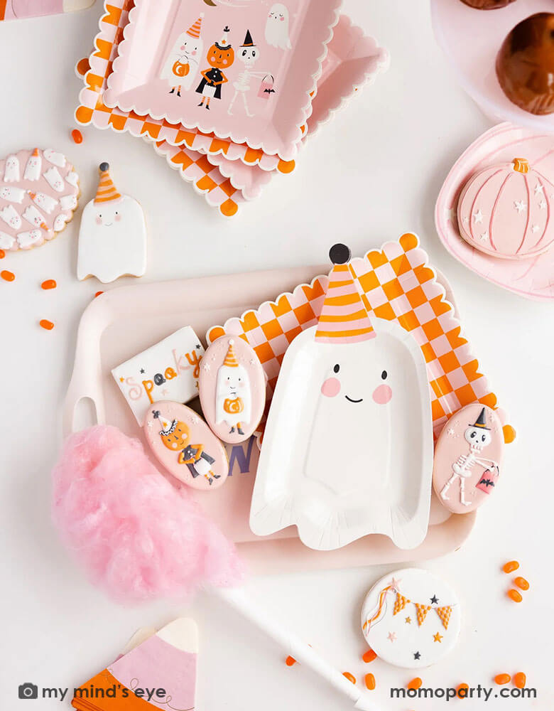 A pink Halloween party table featuring Momo Party's ghoul gang collection by My Mind's Eye including 11.5" x 6.5" ghost shaped plates, orange and pink checkered scallop edged plates, ghoul gang scene paper plates, pink pumpkin plate on a pastel pink reusable bamboo tray. With coordinating decorated sugar cookies, orange jelly beans, pink cotton candy and caramel apples around the table, it makes an adorable pink girly Halloween party inspiration. 