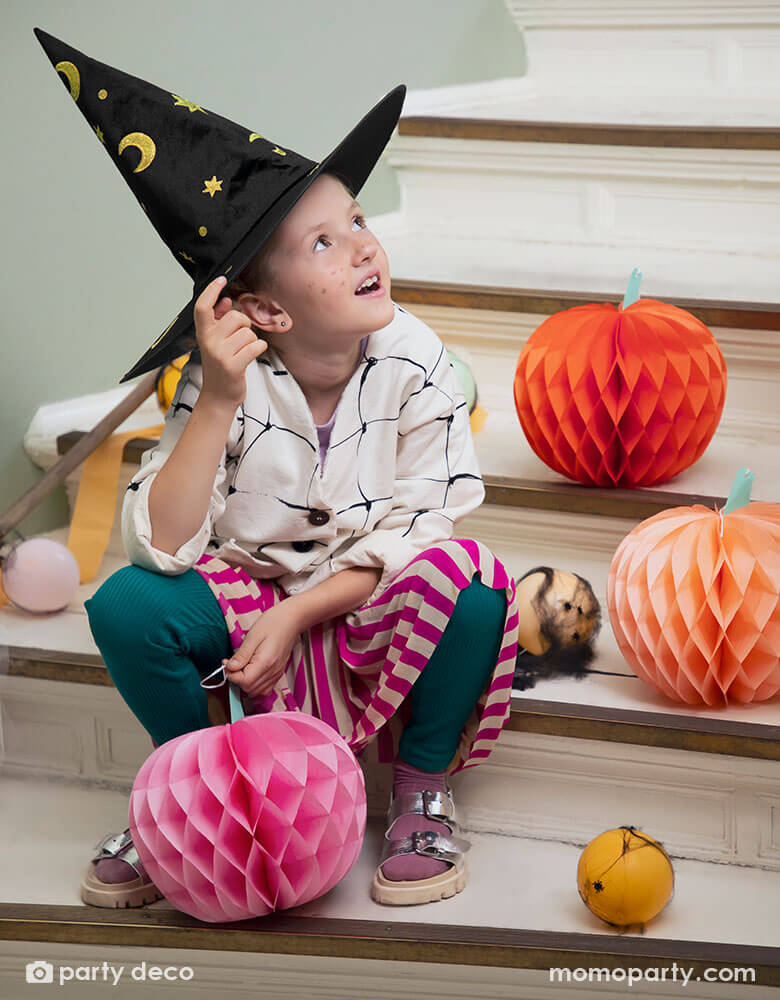 A girl wearing her Halloween costume and a witch hat setting on stairs looking up. Around her there are some party balloons in Halloween colors of orange and pink and blush wrapped in spider web to make a spooky vibe, along with Momo Party's 9.5" pink pumpkin honeycomb set by Party Deco. Comes in a set of 3 honeycomb pumpkins in pink, orange, and peach colors, this set gives a spooktacularly sweet Halloween look.