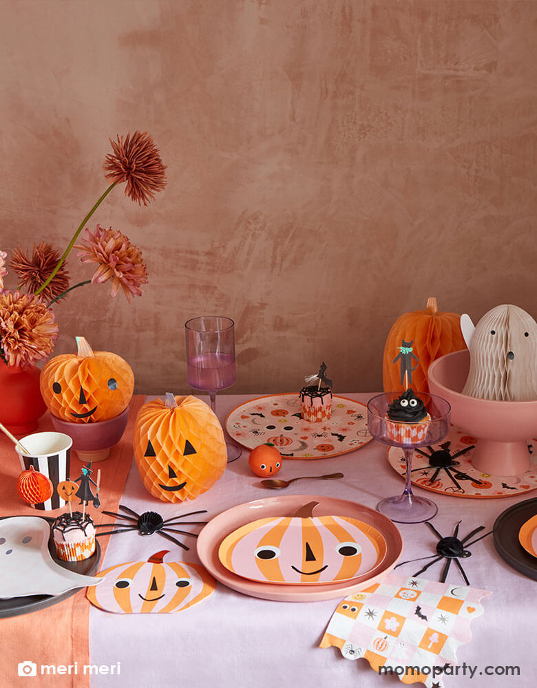 An adorable Pink Halloween party table featuring Momo Party's pink and orange striped pumpkin plates, napkins and groovy Halloween dinner plates & napkins by Meri Meri. On the table there are also cute Halloween honeycomb decorations in the shape of a ghost and pumpkin and spider with spooky cute face on them. With Halloween character cupcakes, it makes a perfect party inspiration for a cute Halloween bash for kids. 