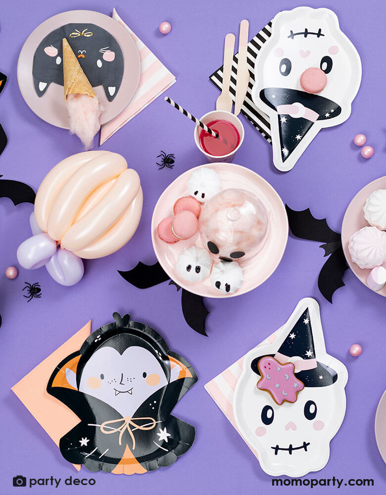 A purple Halloween party table featuring 7" x 10.6" pink skull shaped plate and black cat shaped napkin and pink striped napkin from Momo Party. Around the tableware there are some pink Halloween themed treats including marsh mellows, macaroons, cotton candies, with bat and spider confetti scattered around the table, it makes a great inspiration for a kid-friendly Halloween party.