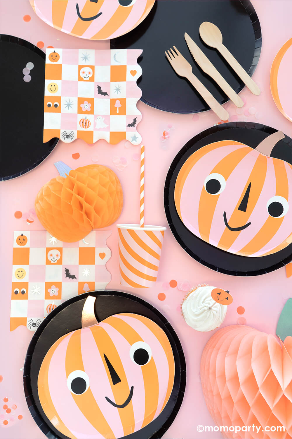 A pink girly Halloween party table featuring Momo Party's orange and pink striped pumpkin shaped plates layered on 10" black large plates, groovy Halloween checkered napkins with Halloween character designs including skulls, pumpkins, bats, spiders ghosts and googly eyes, with honeycomb pumpkins around and festive confetti in orange and pink colors, this makes a great inspiration for a not-so-scary kid-friendly Halloween party tablescape.