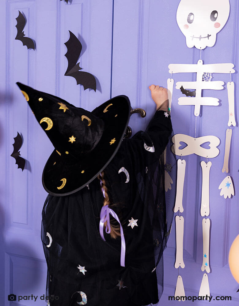 A little girl in witch costume knocking on a purple door for Halloween trick or treat. The door is decorated with Momo Party's skeleton papery decoration and paper bat decorations on it. A spooky fun vibe is in the air this Halloween season!