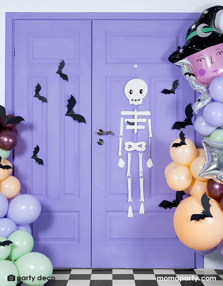A purple door decorated with Halloween themed balloons and decorations including Momo Party's skeleton hanging paper decoration, witch head shaped foil balloon and a Halloween themed balloon garland in lilac, mint, blush and maroon colors, around the balloons there are paper bat decorations to create a spooky vibe a fun Halloween party.