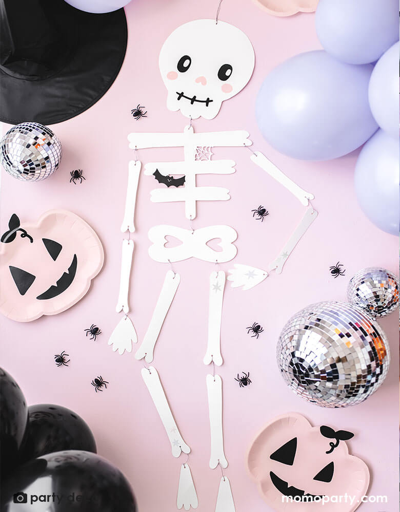 Momo Party's 43" cute friendly skeleton paper decoration on a pink background. Around it is some flittered discos balls, a pink pumpkin shaped plates and spider confetti.
