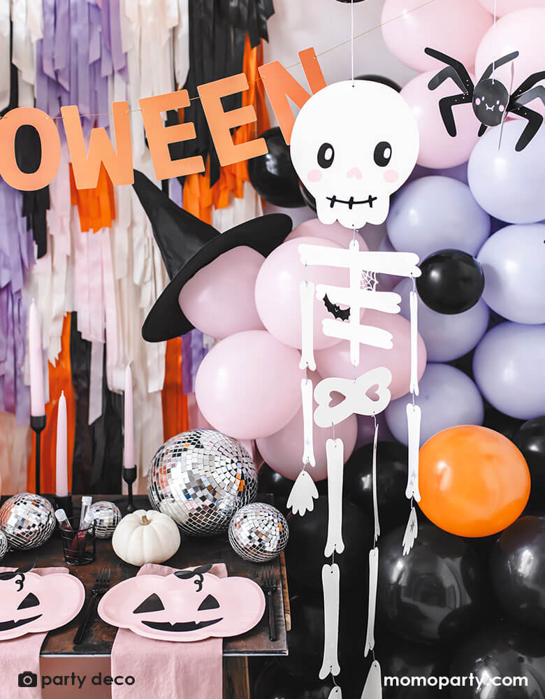 A festive Halloween party set up featuring a beautiful balloon garland in orange, pink, lilac and black, with Momo Party's 14" orange round skull foil balloon, witch's hats, skeleton hanging decoration, paper spider hanging decoration and honeycomb bats in front of a wall of colorful fringe in the Halloween themed colors. In the front there's a party table with some honeycomb pumpkins, disco balls and pink pumpkin shaped plates. All makes a great insp for a kid friendly Halloween bash.