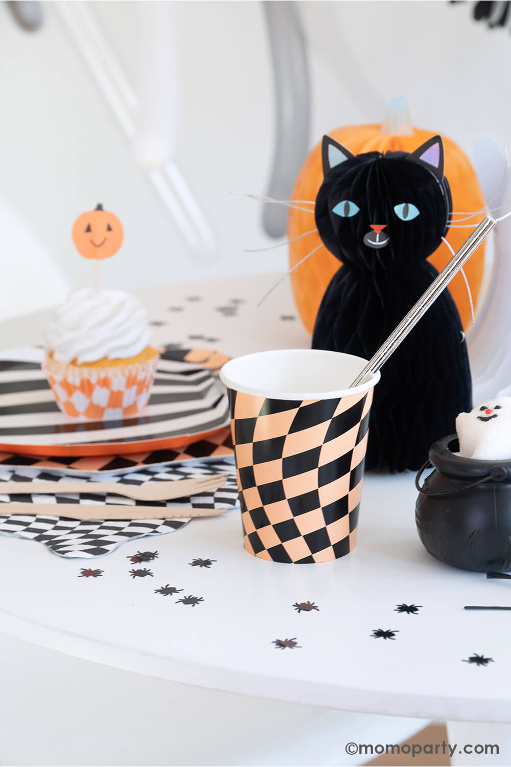 A fun Halloween party table in the classic black and orange colors featuring Momo Party's Halloween Checker Plates, napkins and party Cups by Meri Meri. On the table spider shaped confetti is scattered with honeycomb cat and pumpkin paper decorations as the centerpiece. At the corner there's a mini cauldron container filled with ghost shaped marshmallow treats. All makes a fun and whimsy inspiration for kid's friendly Halloween party ideas.
