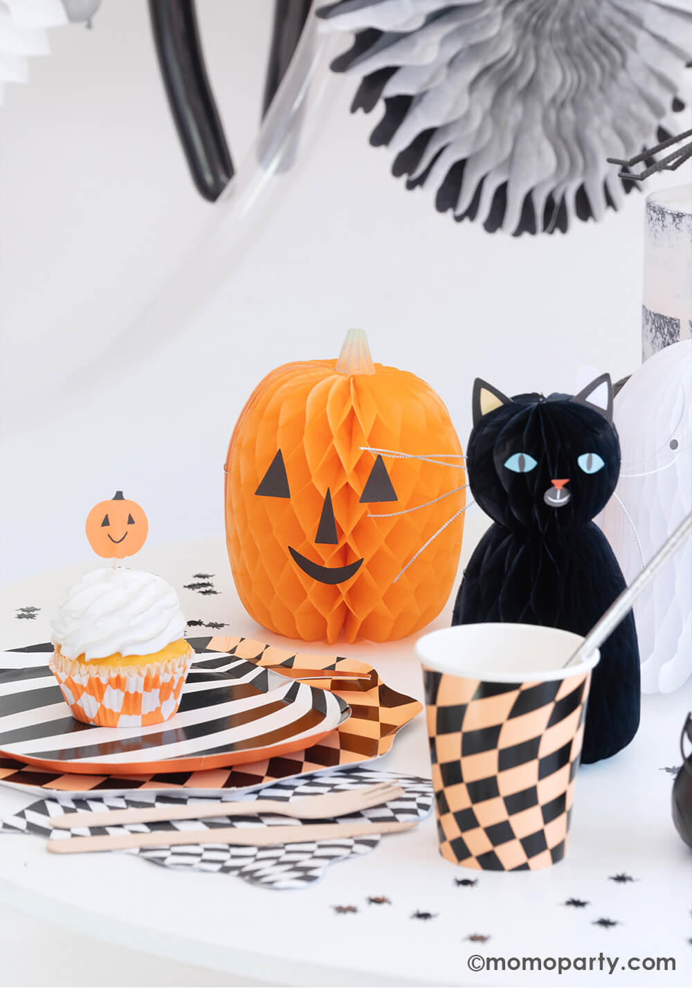 A fun Halloween party table in the classic black and orange colors featuring Momo Party's Halloween Checker Plates, napkins and party Cups by Meri Meri. On the table spider shaped confetti is scattered with honeycomb cat and pumpkin paper decorations as the centerpiece. On the plates there's a cupcake topped with a Halloween pumpkin topper. All makes a fun and whimsy inspiration for kid's friendly Halloween party ideas.