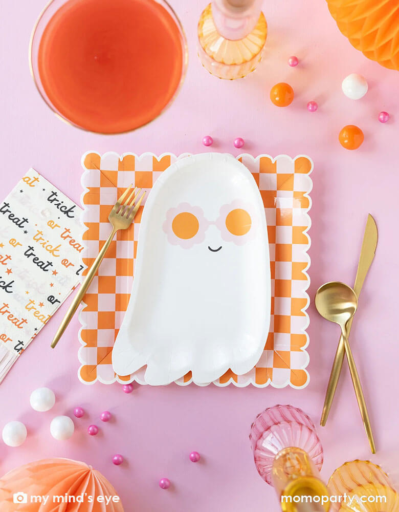 A pink Halloween table featuring Momo Party's pink sunny ghost shaped plates paired with orange/pink checkered plates, with pink trick or treat fringe napkins and honeycomb pumpkins around, it gives a great inspiration for a modern and chic Halloween party table decoration ideas.